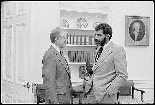 A smiling President Jimmy Carter speaks with journalist Ed Bradley, who has his hands in his pocket.