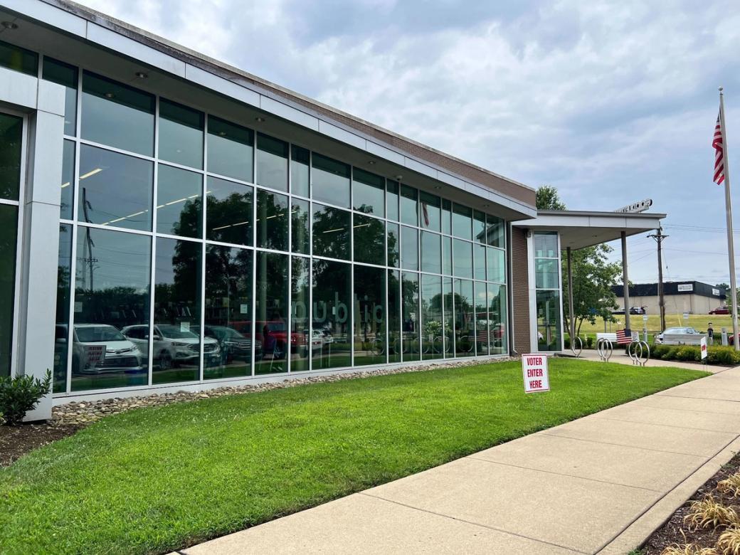 Outside a library with wall-to-wall windows. On a small grassy lot in front of the library there's a white sign that says 'Voter Enter Here' in red letters.