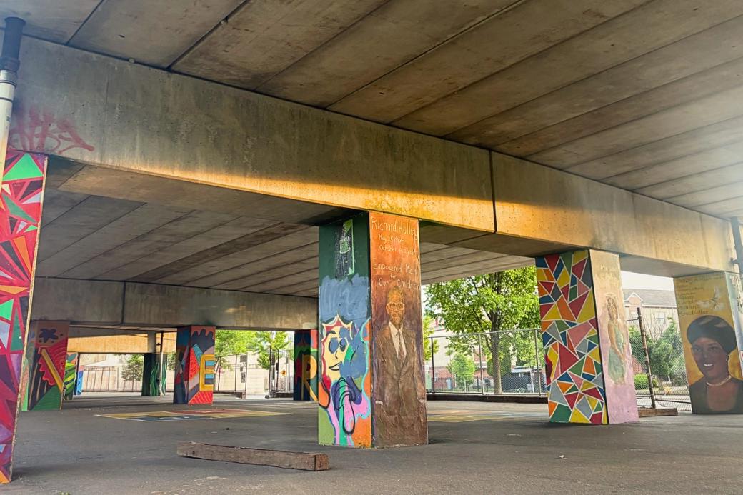 A concrete underpass is covered in colorful murals made golden by the sun starting to set on a warm, sunny day. Green trees are visible past the underpass, and a chain-link fence encloses the space.