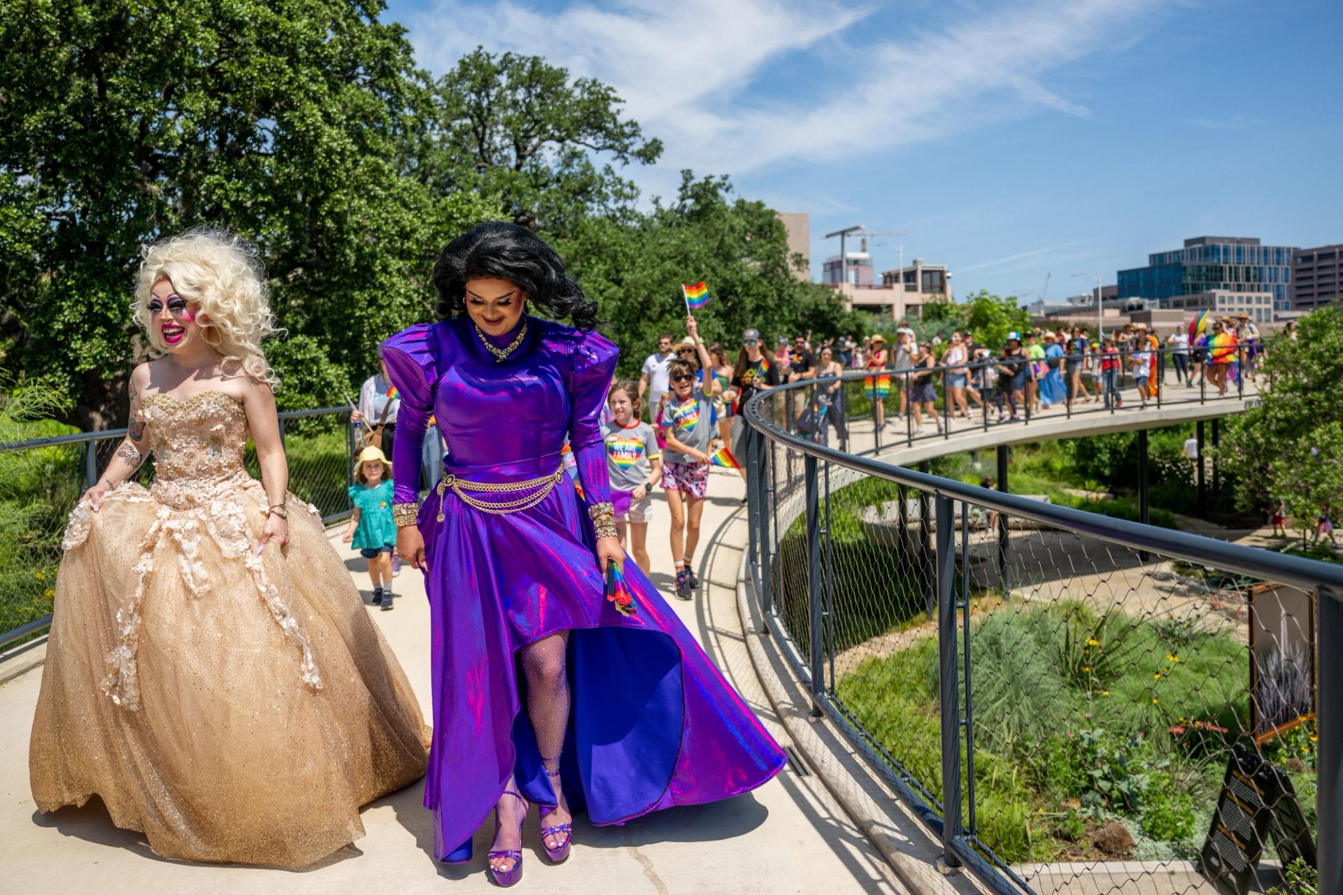 Two drag queens, one in a purple dress and the other in a gold dress, lead a small Pride parade.