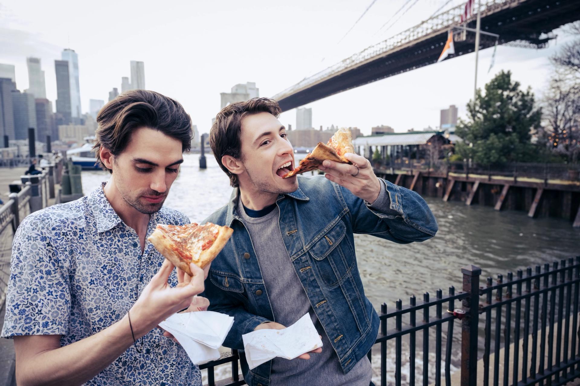 Two men eat slices of pizza near the Brooklyn Bridge in New York City.
