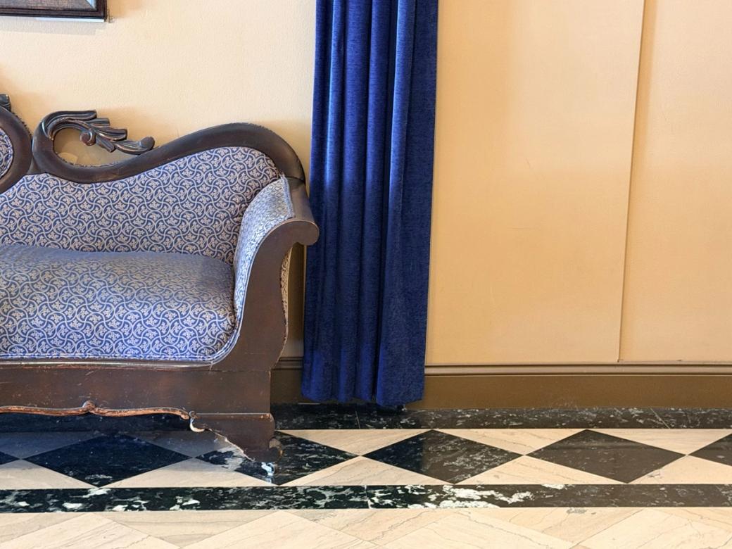 An ornate wooden upholstered couch in blue-and-white patterned fabric sits beside dark blue curtains on a black-and-white checkered marble floor.
