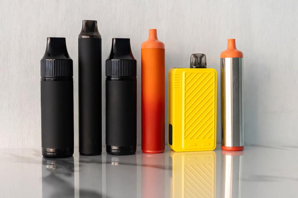 Six e-cigarette products stand in a line, three black, three in bright neon colors of orange and yellow. 