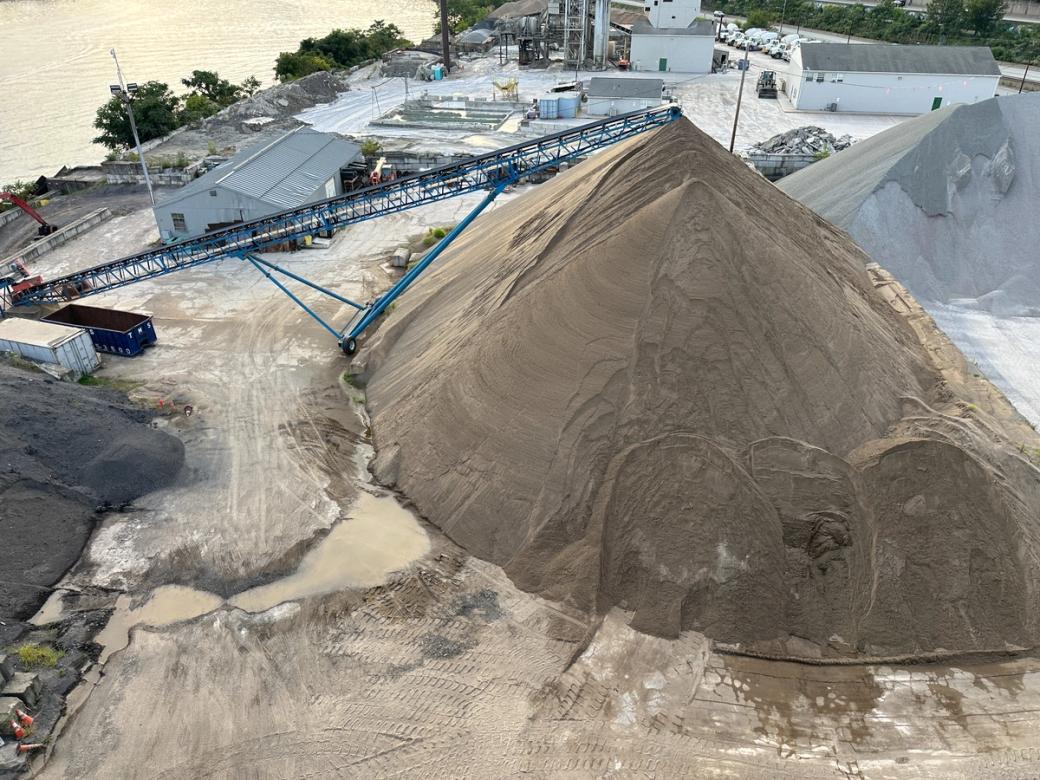  A giant pile of sand that towers over buildings sits near the bank of a muddy river.