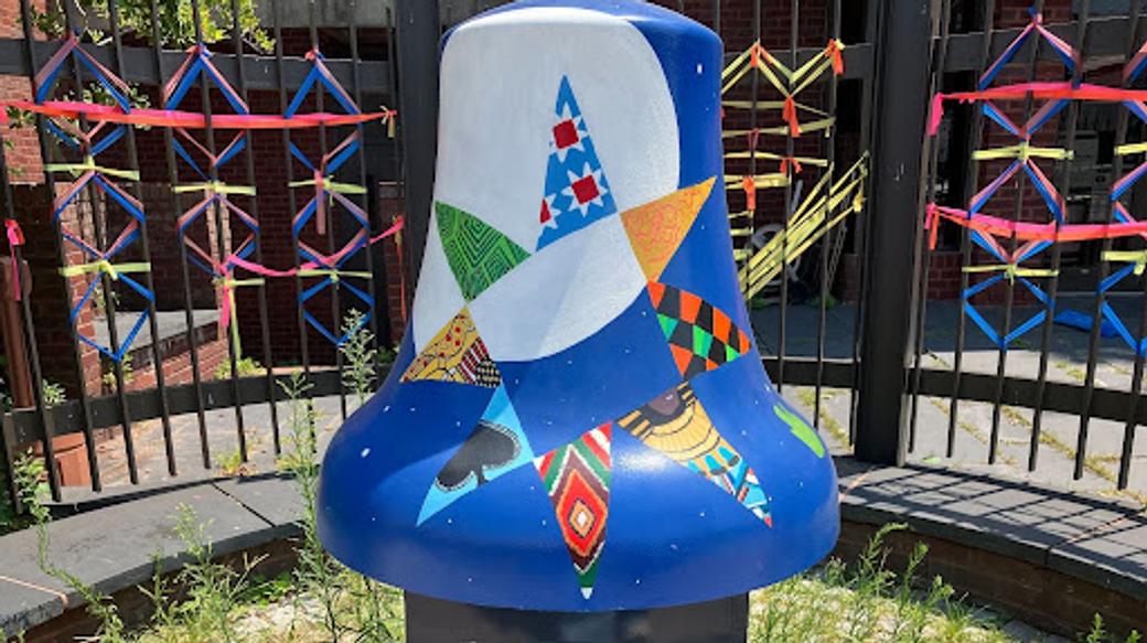 A large blue and white bell sits on a pedestal outside a gated building. At the center of the bell is a star with each pointed painted in a different multi-colored pattern. Colorful string is tied around the gate.