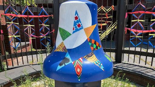 A large blue and white bell sits on a pedestal outside a gated building. At the center of the bell is a star with each pointed painted in a different multi-colored pattern. Colorful string is tied around the gate.
