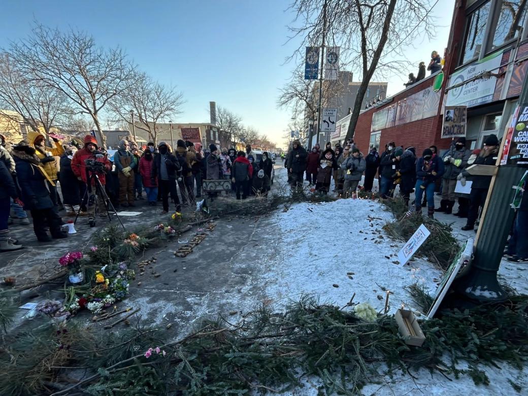Group of adults stand around memorial on sidewalk in winter, snow covers much of the ground