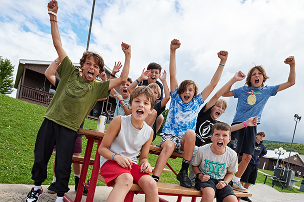 A group of excited kids sit and stand on a picnic table, cheering with raised arms. They display joyful expressions, set against a cloudy sky and grassy area.