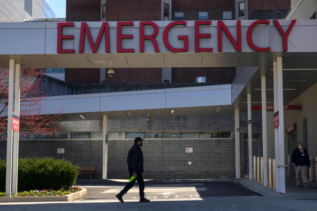A person walks beneath a large red “Emergency” sign at the entrance of Penn Presbyterian Medical Center. A person in a mask walks away from the emergency room entrance.