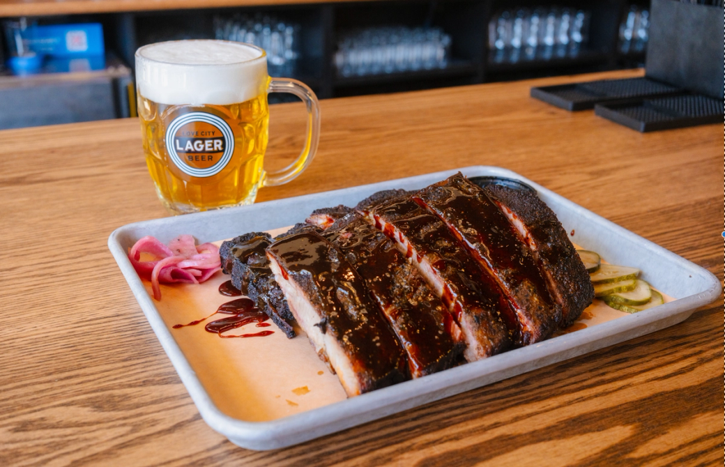 Barbecued ribs smothered in a dark red sauce sit on a grey tray, with pickles and pickled red onions lining the sides. The meal sits on a wood counter with a golden Love City Lager beer topped with a substantial beer foam to the left. 