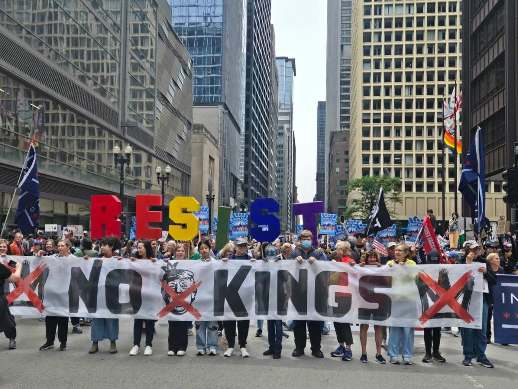 Protest march on city street, with participants holding signs that say "RESIST" and "NO KINGS"