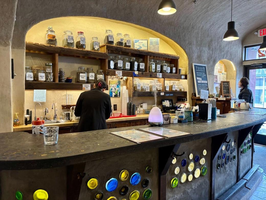 A person stands behind a concrete bar with colorful circle tiles. Jars of tea on the wall.