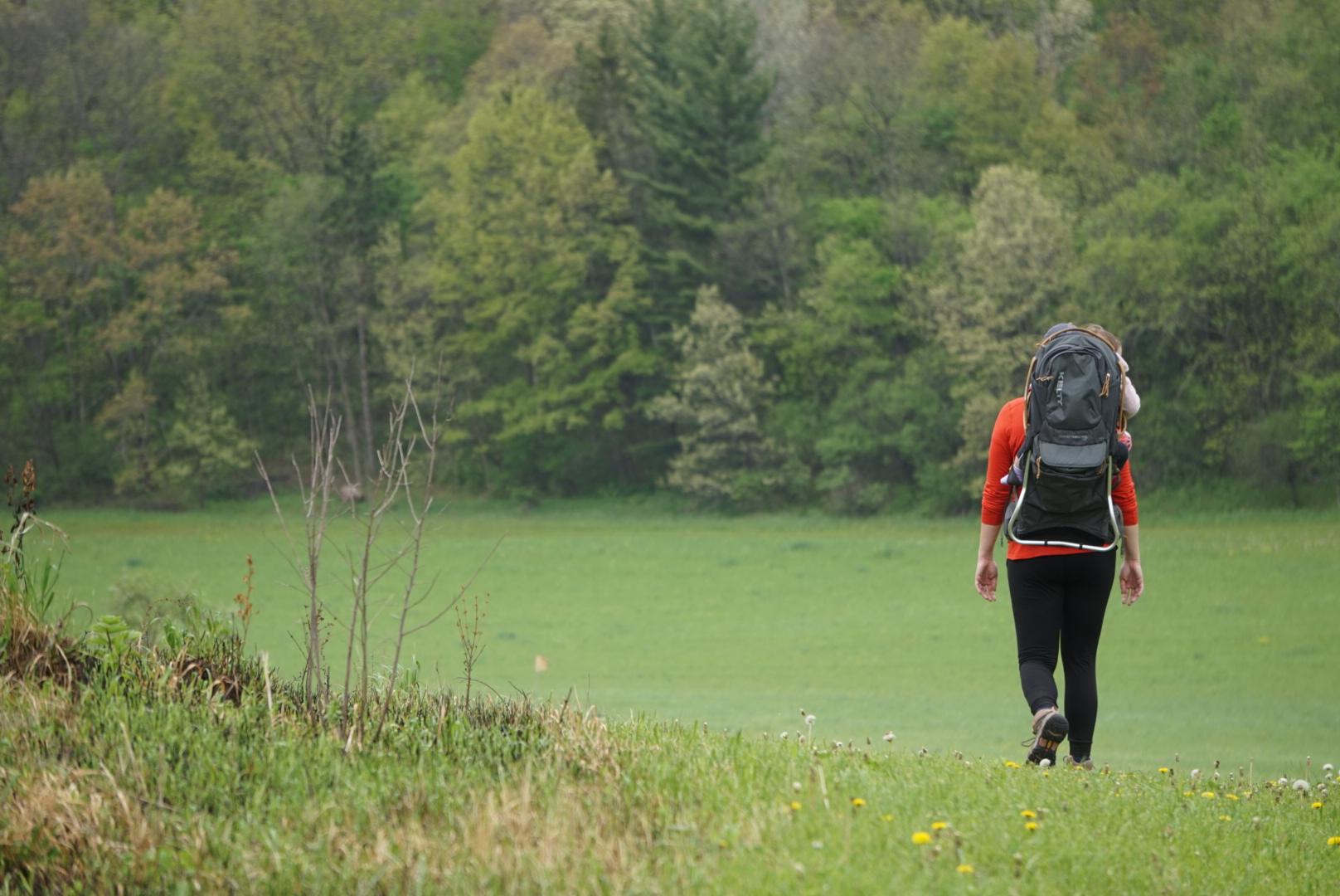A woman in a backpack and red shirt walks across a field.