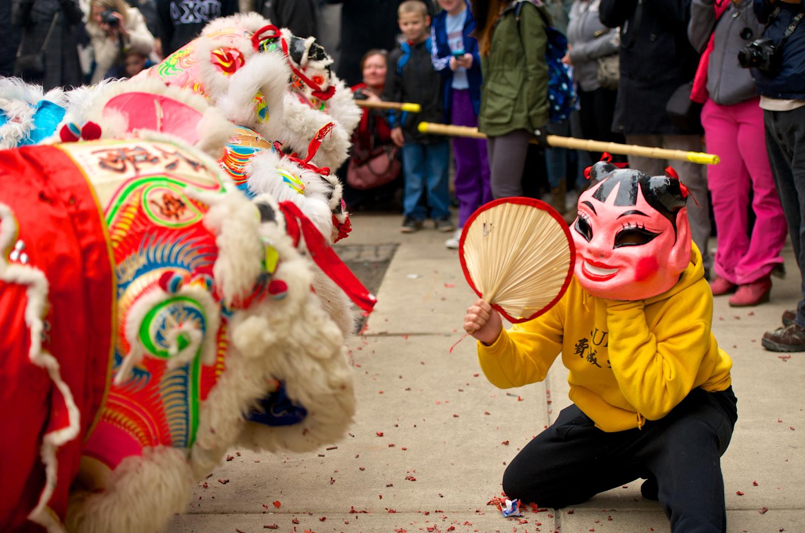 A person in a colorful mask with a fan kneeling on the ground.
