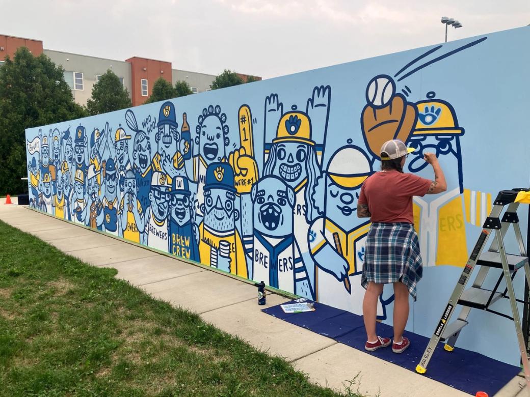 A woman paints a mural on a wall. The wall is painted cornflower blue and is populated with people and animals wearing Brewers uniforms and caps.