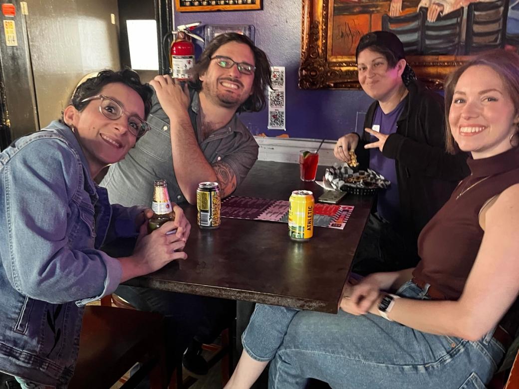 Four people sit at a wooden table drinking alcoholic beverage, and smiling at the camera.