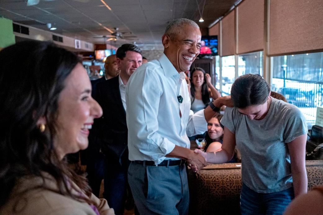 Former President Barack Obama smiling while chatting with people in a restaurant.