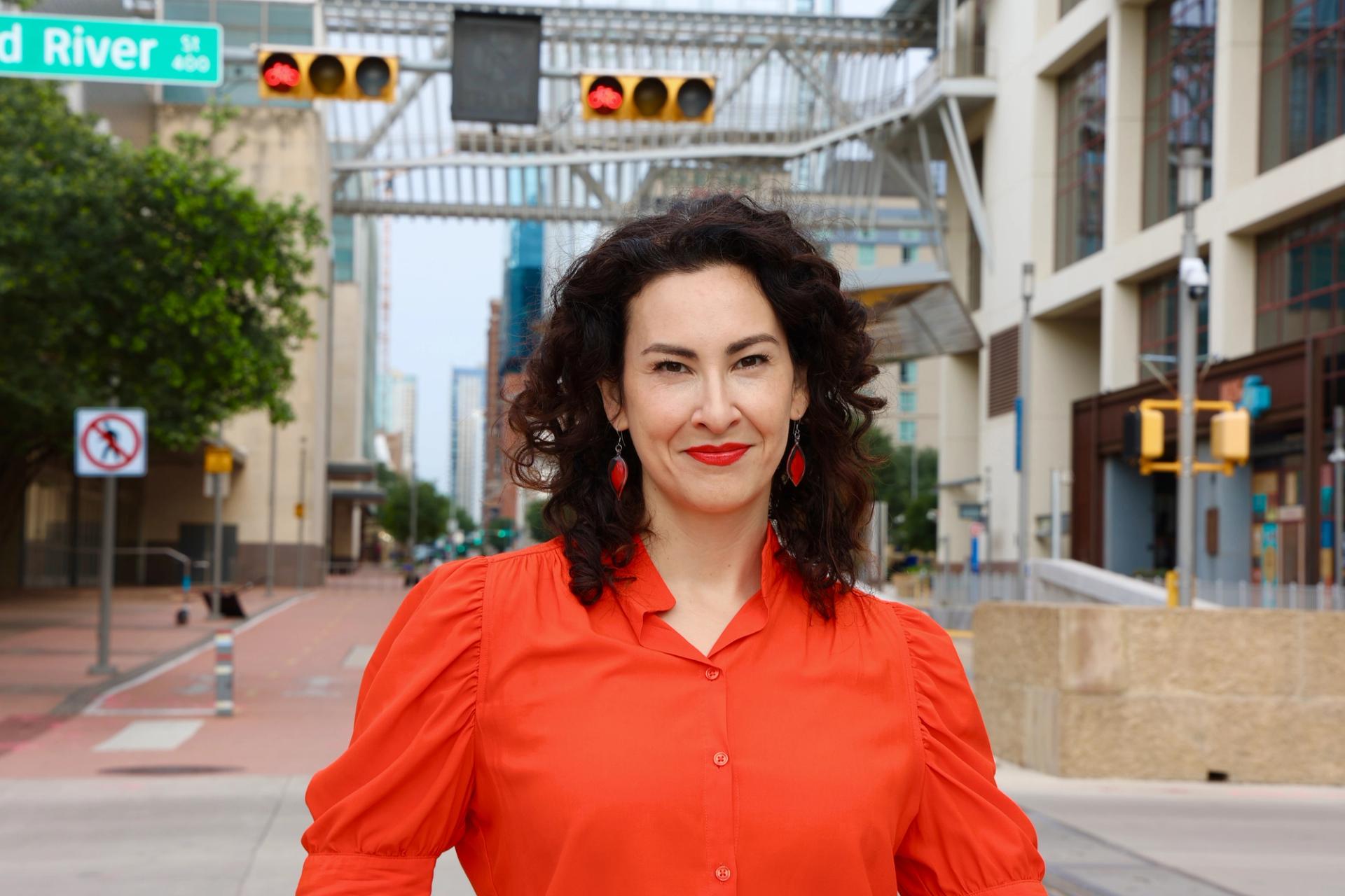 A woman with dark curly hair wearing a bright red shirt on a road. She is wearing red lipstick and red earrings.