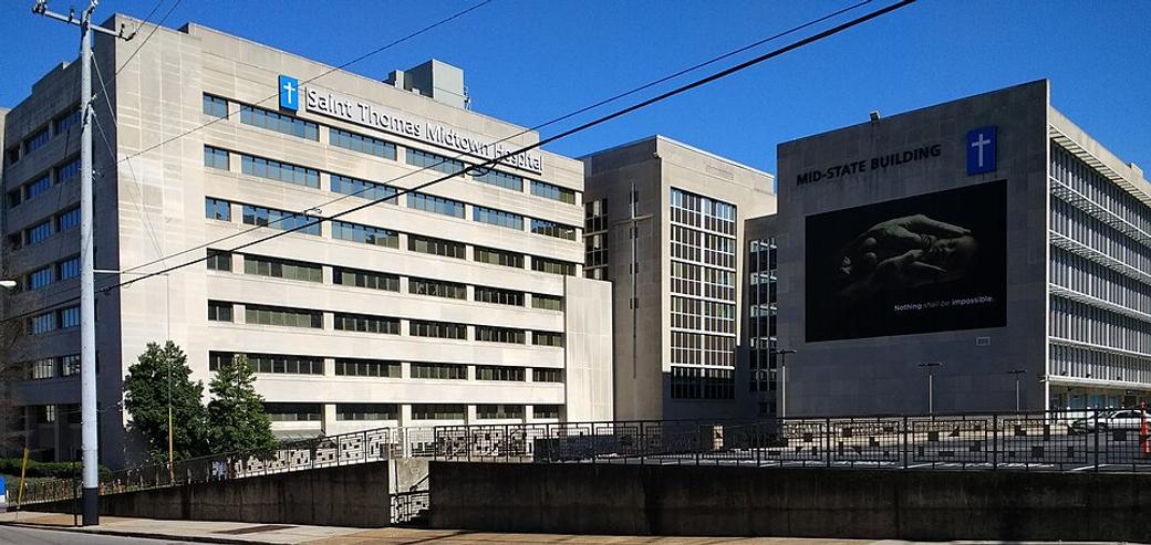 A large gray hospital with a blue sign with a white cross in the center, and the words Saint Thomas Midtown Hospital in white lettering.