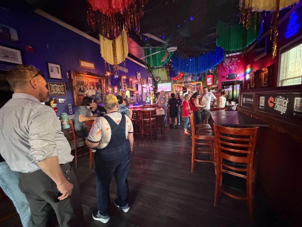 Dimly lit, colorfully decorated bar interior with patrons gathered at tables and a bar, fringe streamers hanging from the ceiling, and two people in the foreground facing away from the camera.