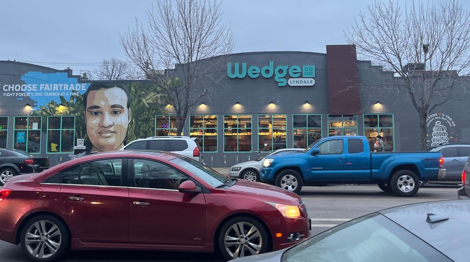 A view of a storefront from the parking lot. On the left side there's a mural with a man in front of brush. To the murals left it reads "choos fairtrade. fight poverty one farmer at a time." On the right it reads "wedge co-op Lyndale"