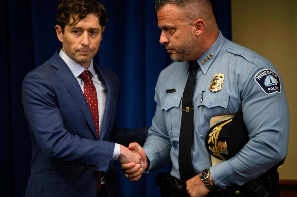 Two men shaking hands on a stage. The man on the left is wearing a suit and tie, the man on the right is in a decorated police uniform