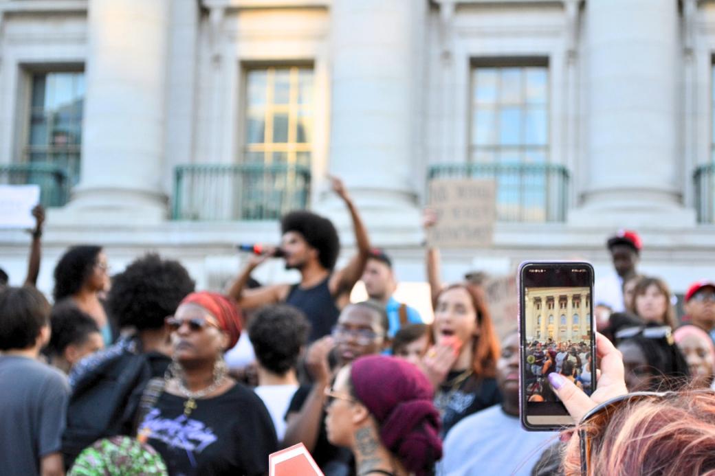 The focus is on an individual taking a photo of protests occurring in front of the Capitol Building. The protestors' faces are out of focus. 