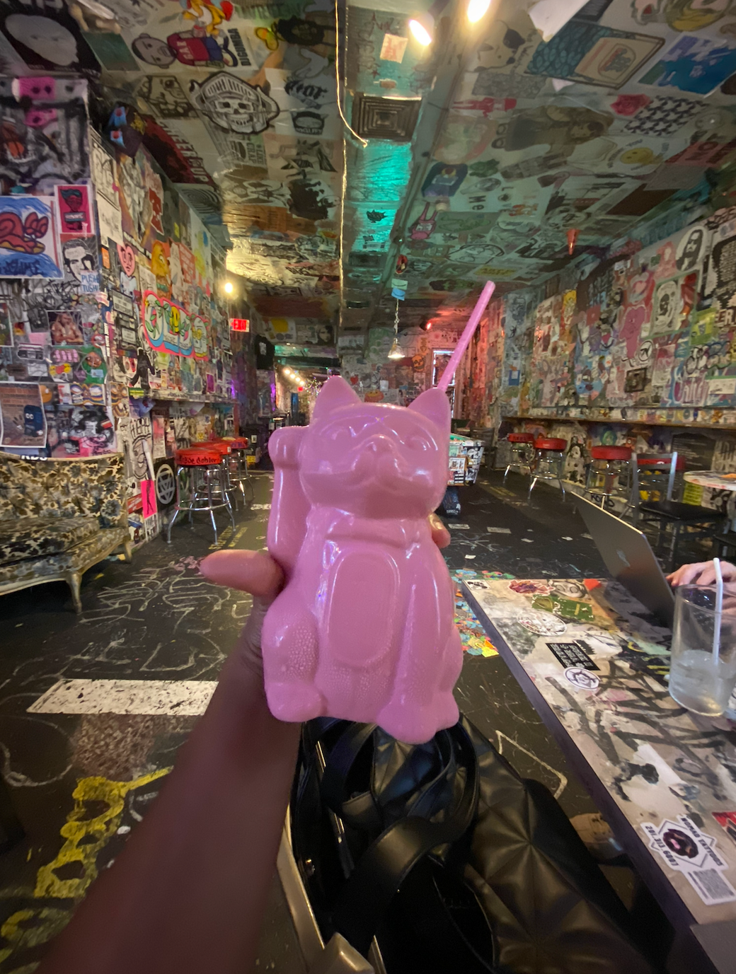 holding a pink kitten shaped glass with a pink straw sticking out of it inside a bar plastered with art and posters and graffiti