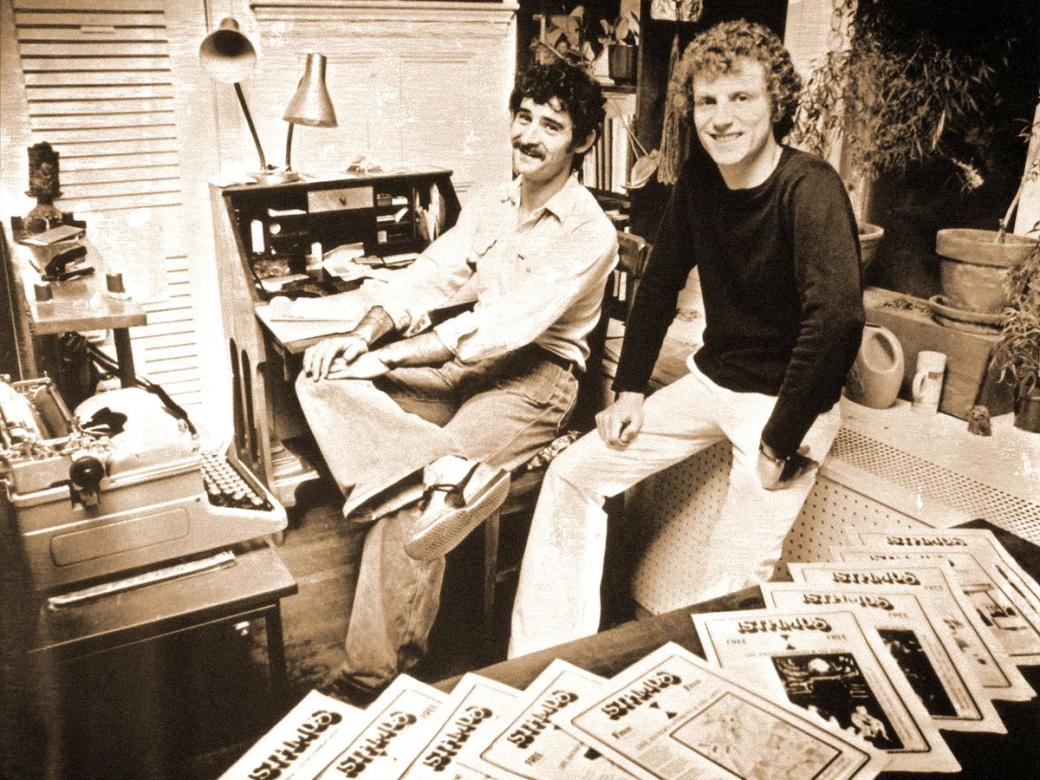 A black-and-white photo of two men surrounded by newspapers.
