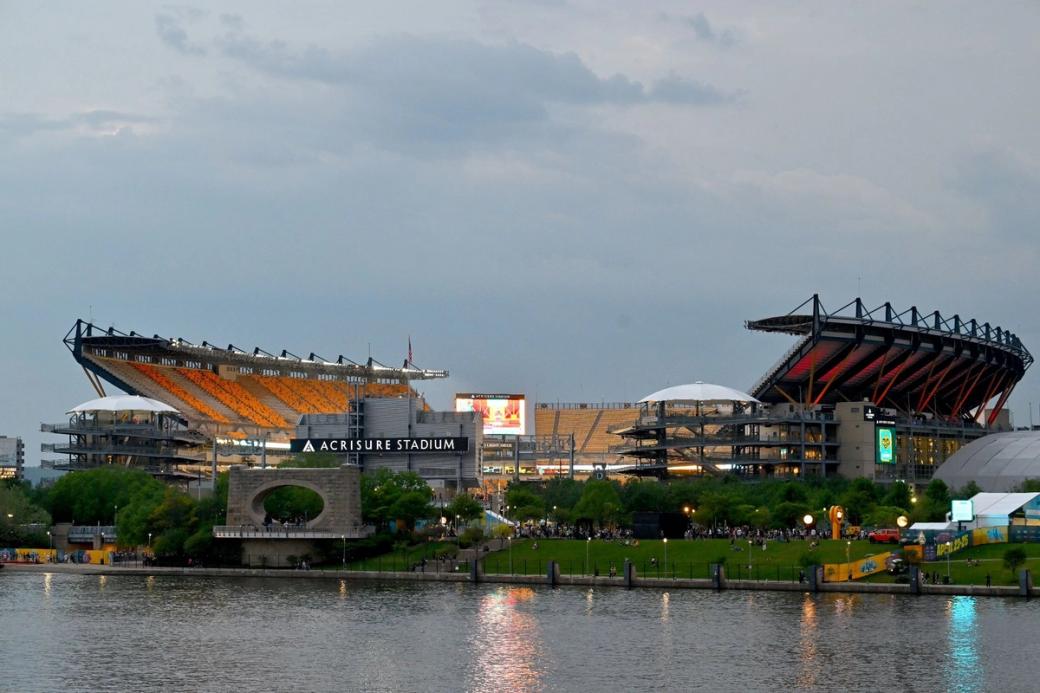 Acrisure Stadium lit up from across the river under a cloudy sky.