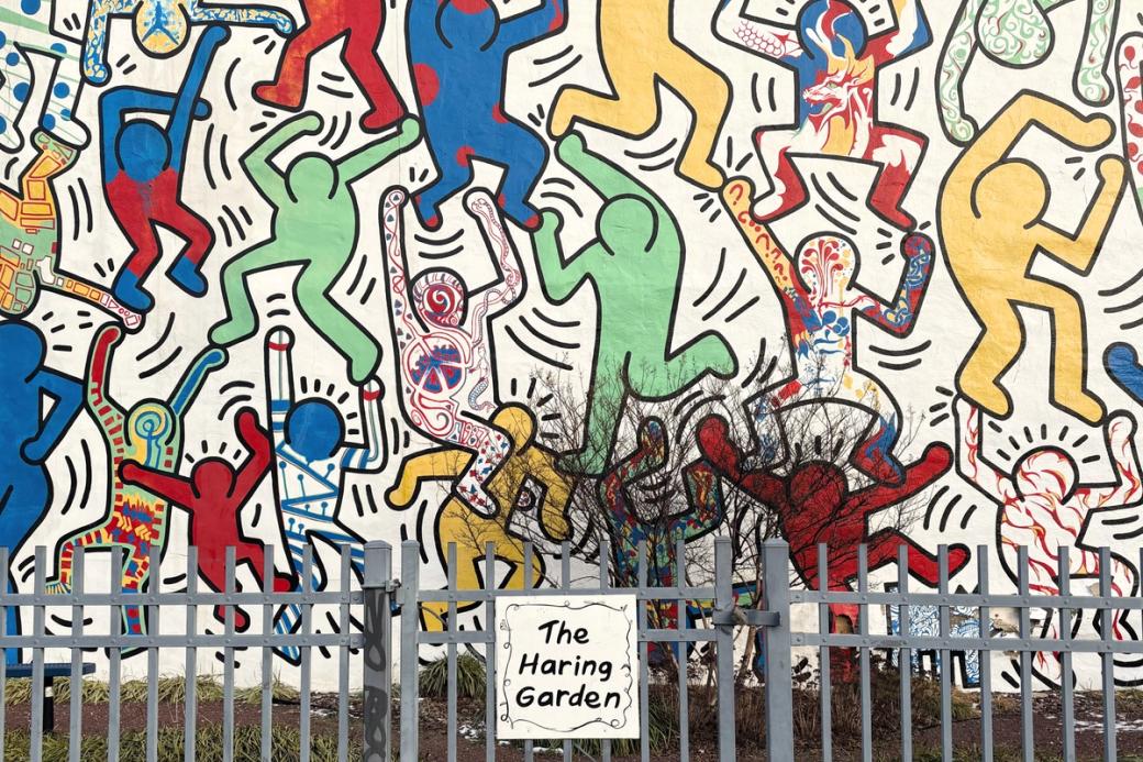 A white sign with "The Haring Garden" written in black graces the front of a gray gate. The small garden houses the Keith Haring mural “We The Youth,” which features his iconic black-outlined figures filled in with bold colors and patterns created by local youth. 