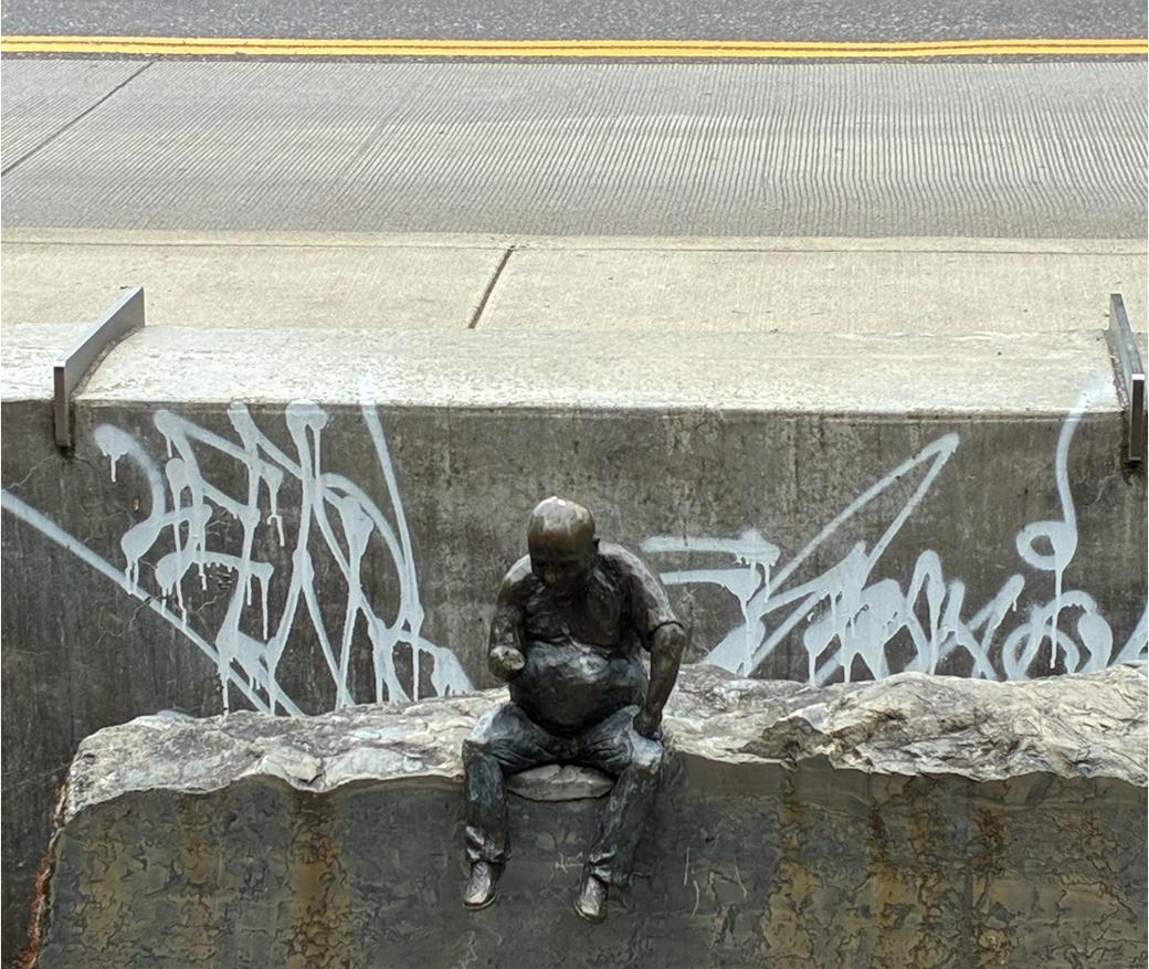 Small sculpture of a figure of a man on rough stone. White graffiti behind him and, behind that, the sidewalk and street.