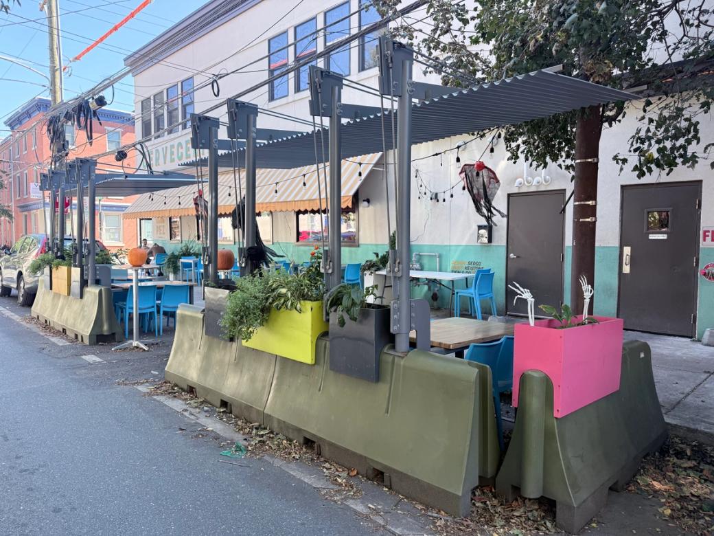 An outdoor streetery protected by concrete barriers. Halloween decorations decorate the display and colorful plant boxes.