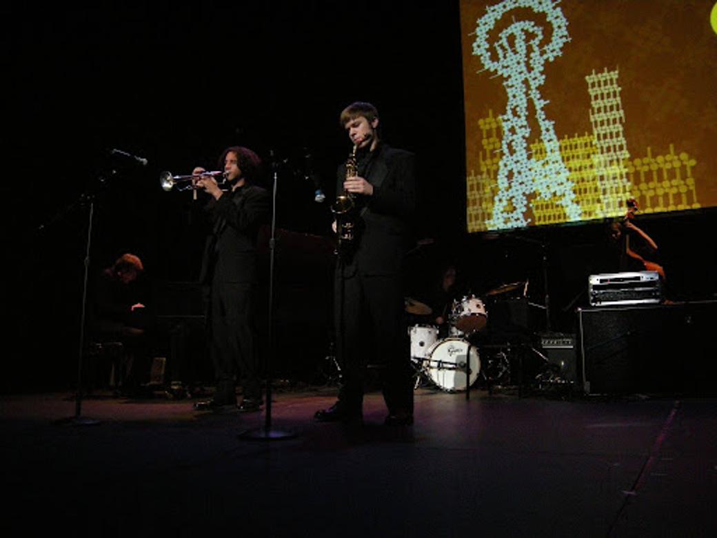 Two Garfield High School students play brass instruments on a darkly lit stage with an artistic rendering of the Space Needle projected behind them.