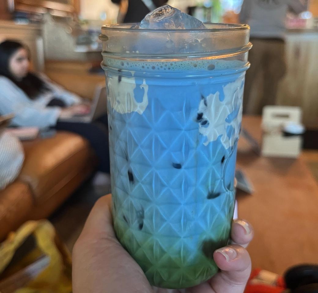 A hand holds a mason jar-style glass filled with a blue-to-green ombré matcha drink topped with an ice cube, in a cafe with people seated in the background.