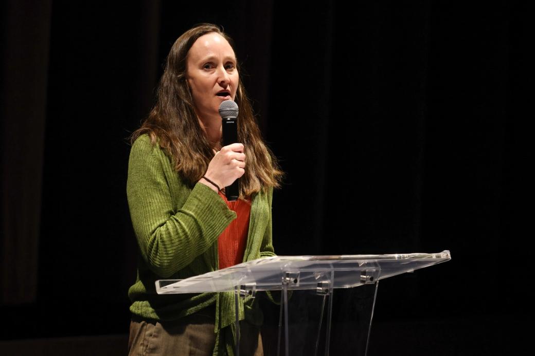 A white woman with brown hair in a green cardigan stands at a podium holding a microphone