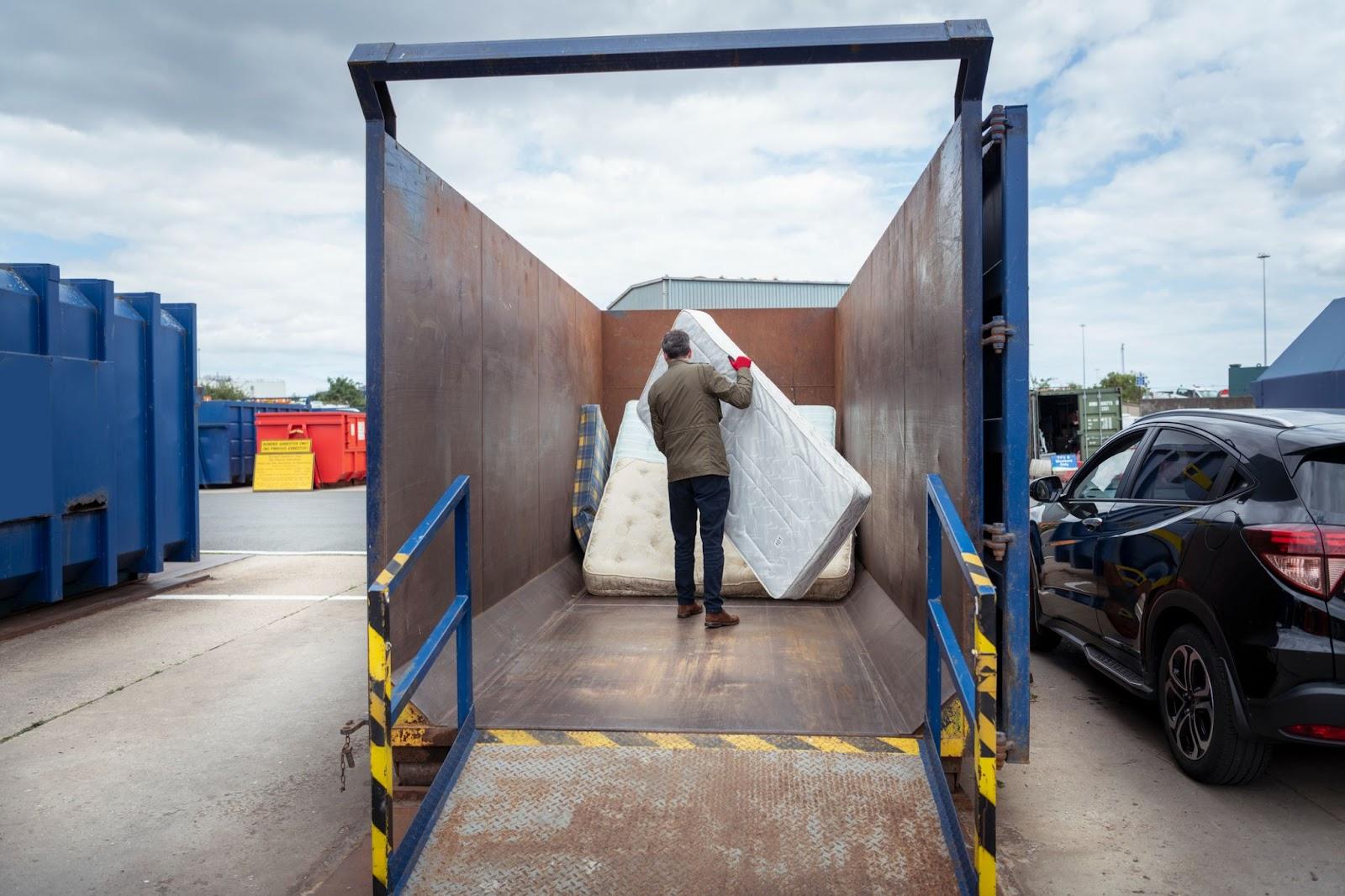 Person walks a mattress into a giant waste container.