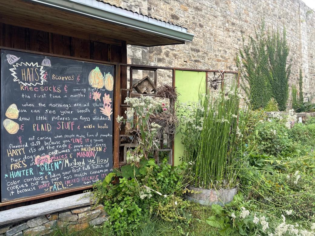 A black chalkboard is covered with fall clothing recommendations in an outdoor garden. Green plants line the bottom border and right side of the photo, and a tall stone wall is behind the board. Dark green ivy crawls up the wall on the right side. 