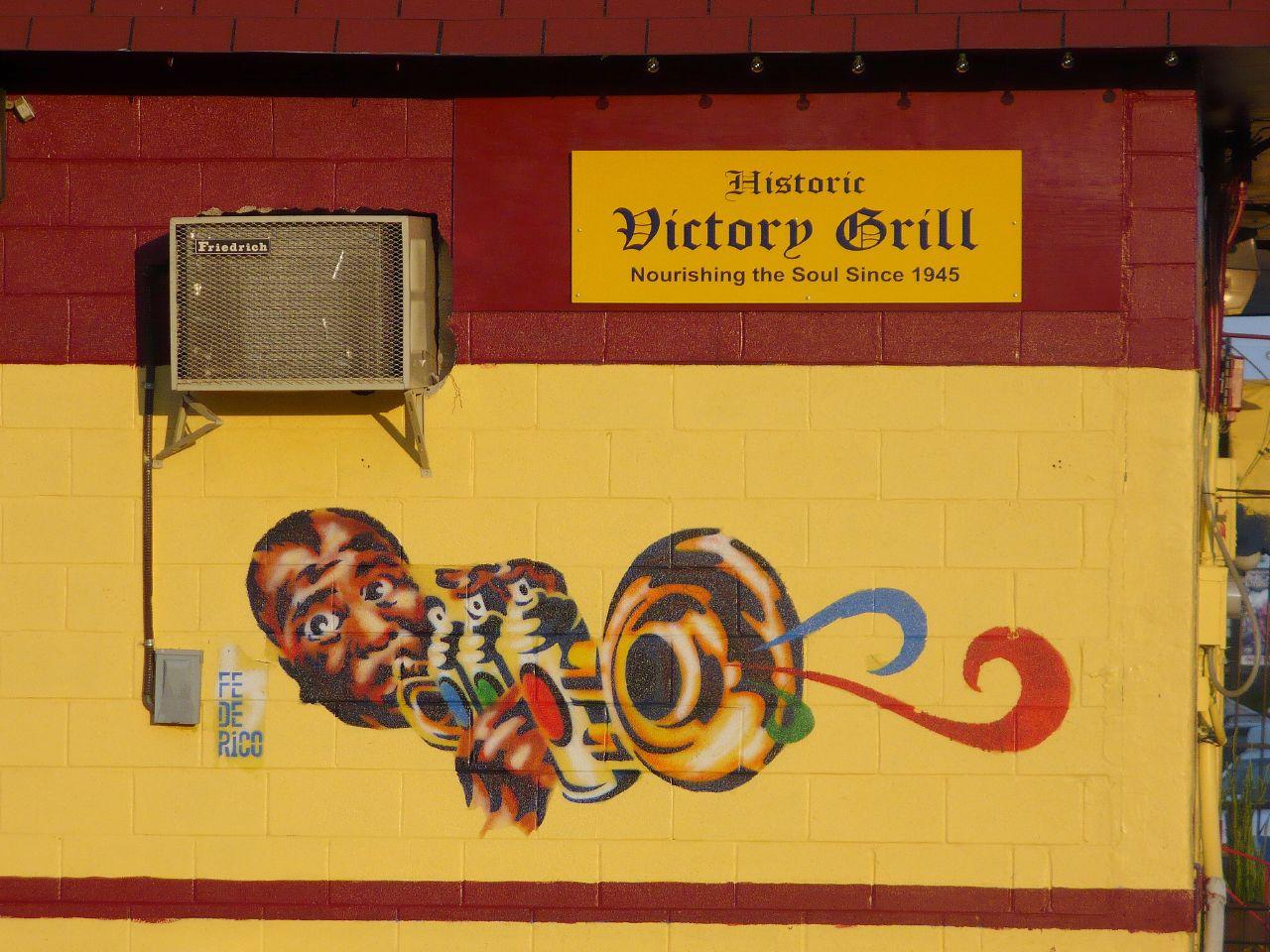 The outside of Victory Grill. A yellow wall has a mural of a man playing the trumpet.