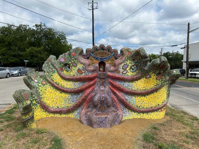 A piece of art shaped like what appears to be a bug and created with colorful mosaic tiles sits on a grass patch in the middle of a road.