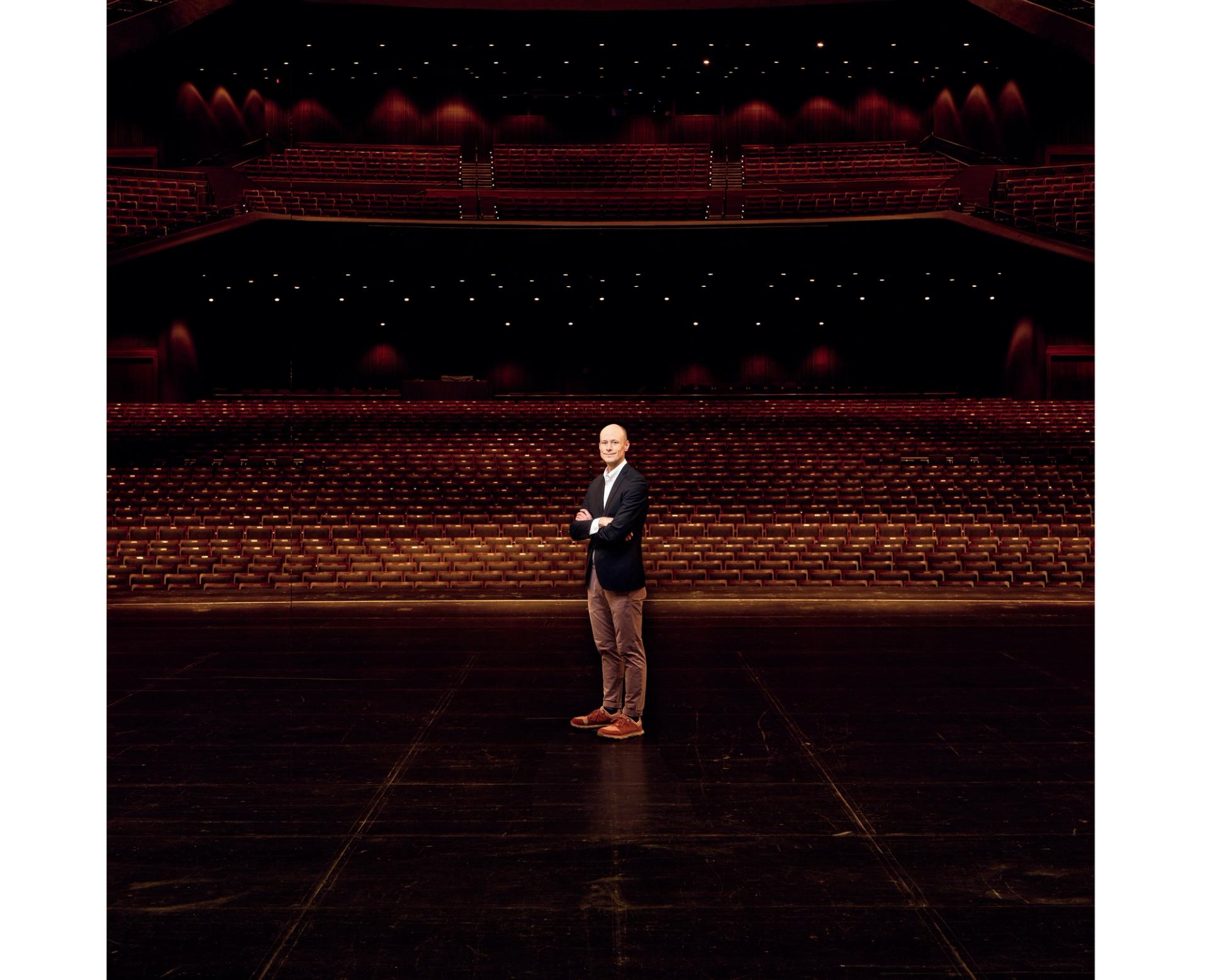 A bald man wearing brown pants, brown shoes, white shirt, and a navy suit jacket standing on a stage in a theater. The audience seats are behind him.