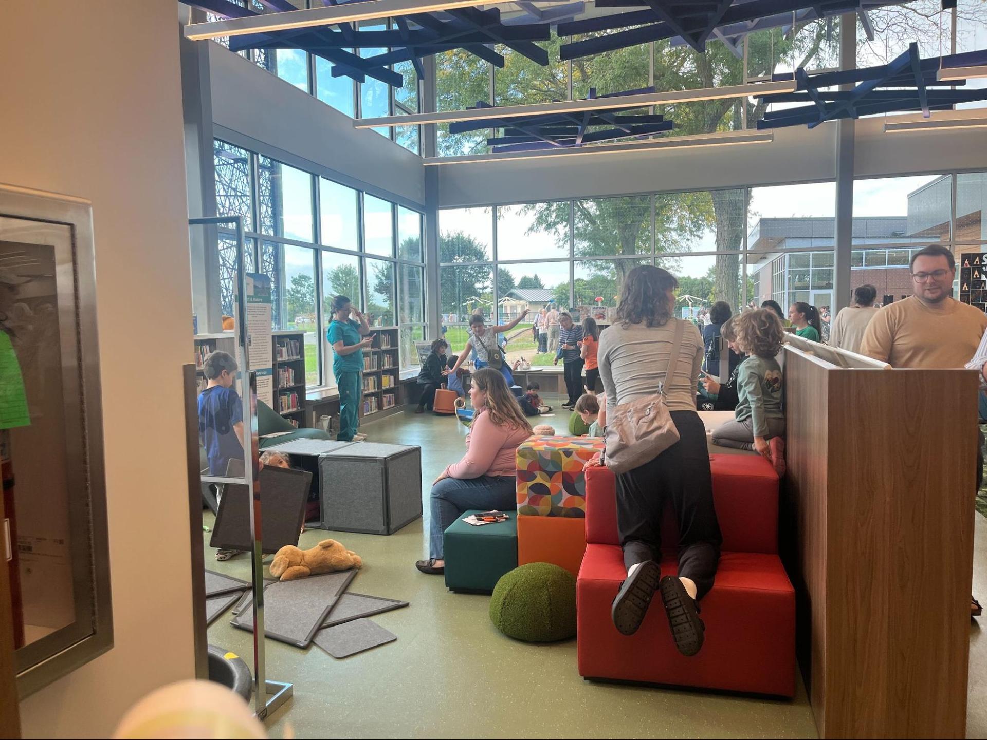 Floor to ceiling windows line the walls of the Imagination Center's children's book area. Several children and parents look at books on shelves and listen to a reading.