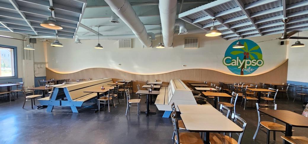 A brightly lit restaurant with light brown tables and benches. A turquoise sign with a palm tree on it says Calypso Cafe.
