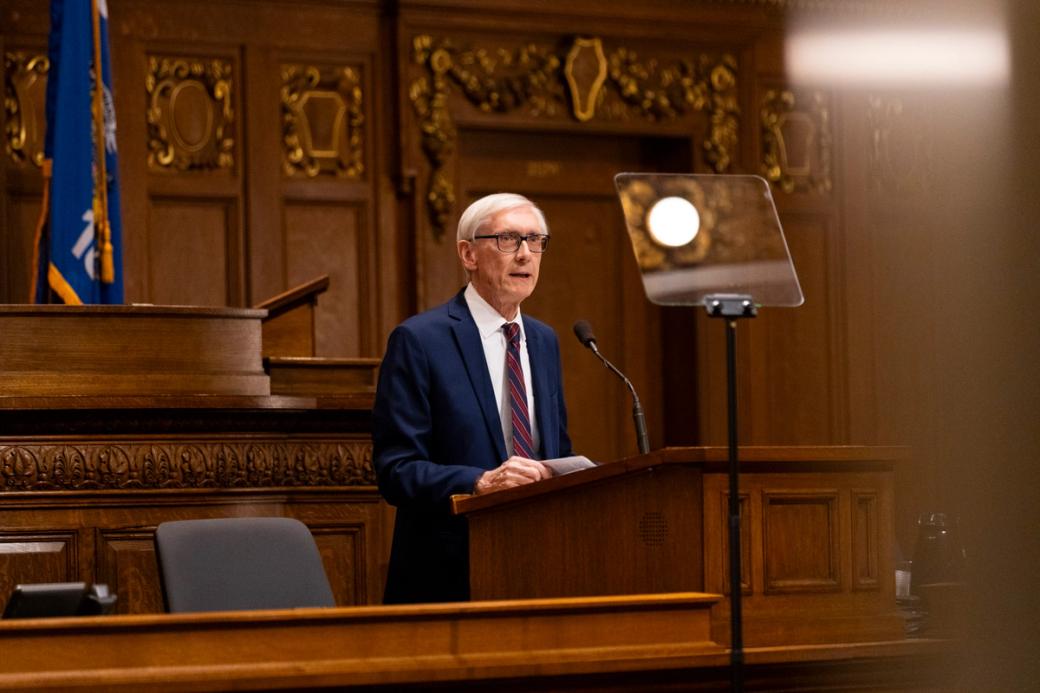 A white-haired man at a suit speaks at a podium.