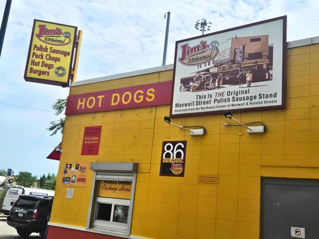 A yellow buildingthat says "hot dogs" There is a sign that says "Jim's Original Polish sausages, pork chops, hot dogs, burgers"