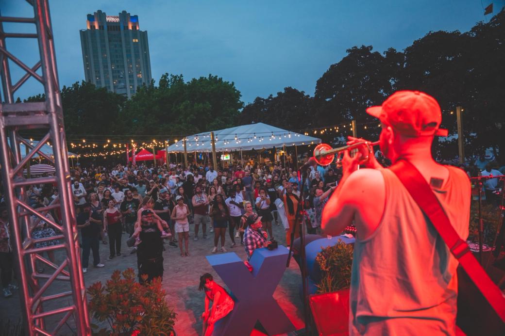 A trumpet player performs on stage in front of a crowd at Spruce Street Harbor Park. String lights hang above the crowd, and the Hilton Hotel is visible in the background. 