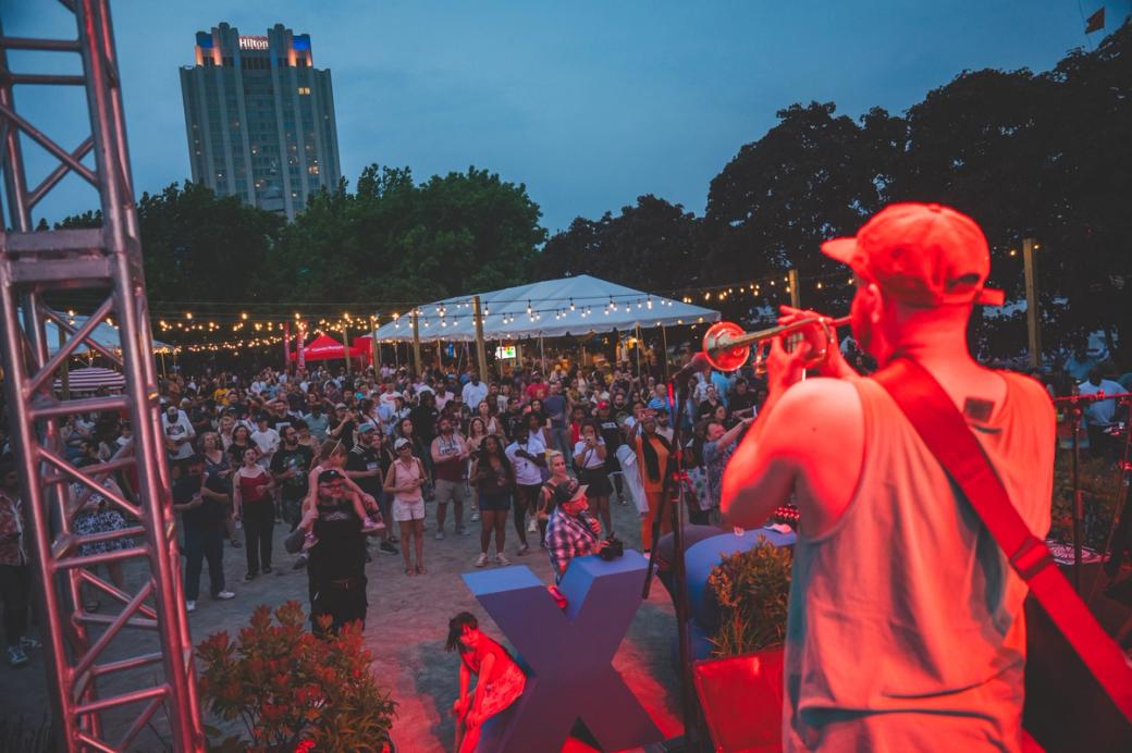 A trumpet player performs on stage in front of a crowd at Spruce Street Harbor Park. String lights hang above the crowd, and the Hilton Hotel is visible in the background.