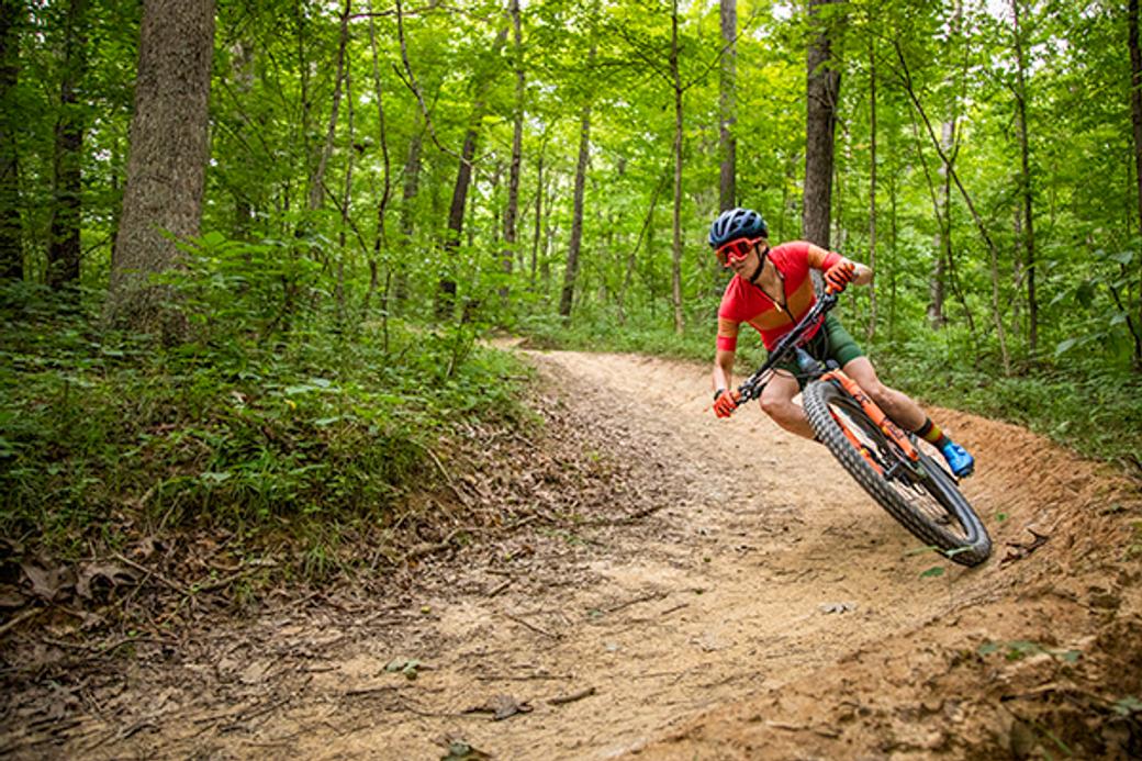 Mountain biker in a red shirt leans into a curve on a forest trail. Lush green trees surround the path, creating a sense of adventure and focus.