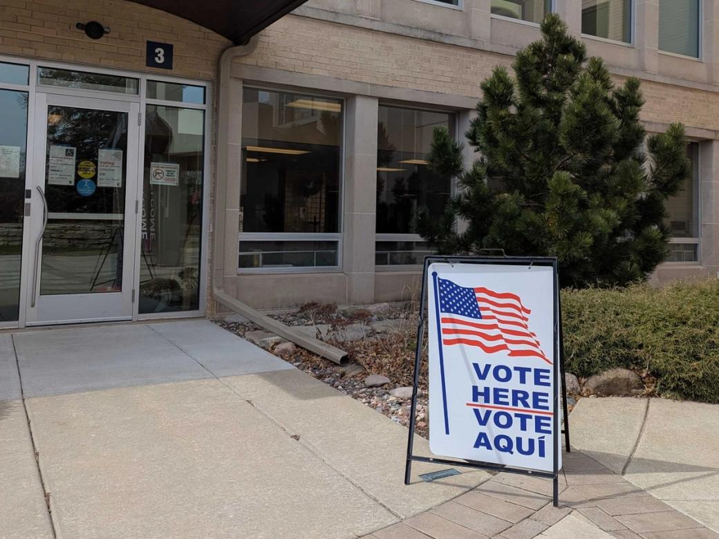 A white sign with a United States flag image and the words “VOTE HERE VOTE AQUI” stands upright in front of a Madison municipal building.