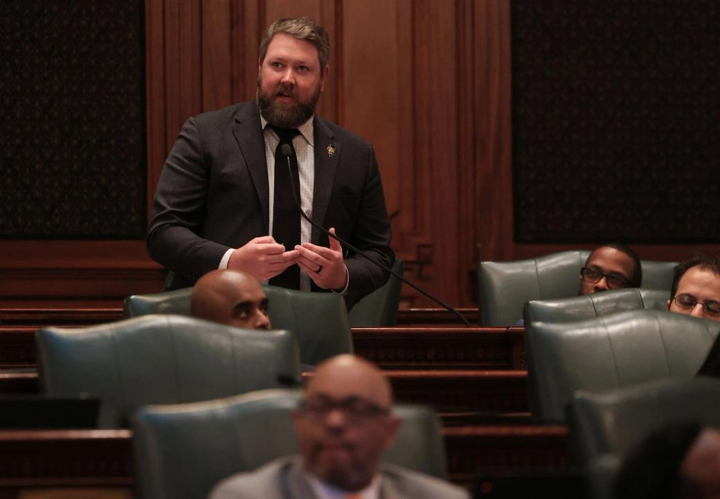 A white man in a suit standing among others seated, in a wood-paneled room.